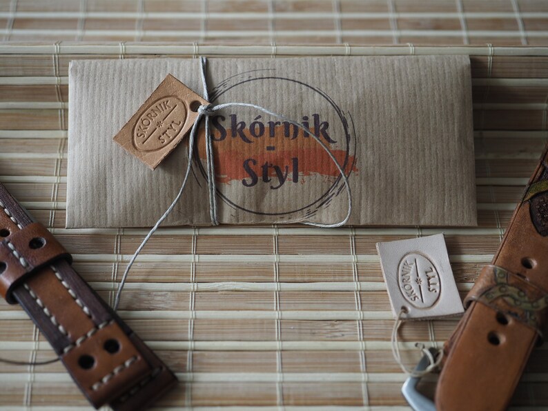 May include: A brown leather watch strap with stitching and two holes. The strap is packaged in a brown paper envelope with a string tied around it. The envelope has a brown leather tag with the text "Sk&oacute;rnik - Styl" on it.