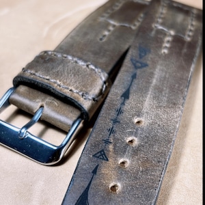 May include: Close-up of a distressed brown leather watch strap with a silver buckle. The strap features decorative stitching and arrow designs. The leather has a worn, vintage appearance, with several adjustment holes.