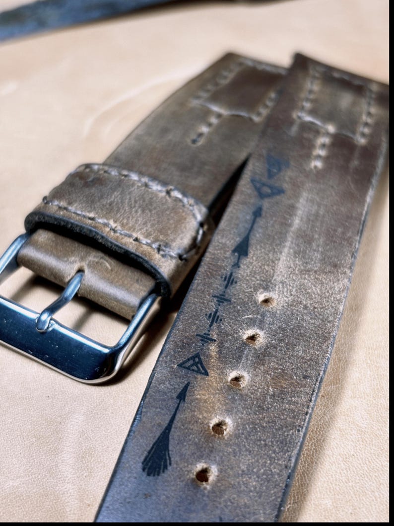 May include: Close-up of a distressed brown leather watch strap with a silver buckle. The strap features decorative stitching and arrow designs. The leather has a worn, vintage appearance, with several adjustment holes.