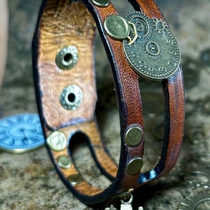 May include: Brown leather bracelet with a steampunk-style clock face and brass rivets. The bracelet features a double-strap design and a small camera charm. The leather has a distressed look, and the metal accents add a vintage feel.