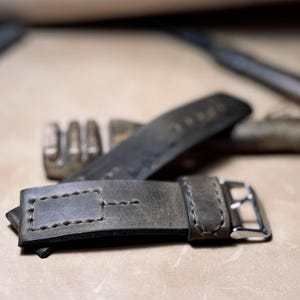 May include: Close-up of a grey leather watch strap with a silver buckle. The strap has visible stitching and a rectangular loop. Another strap is visible in the background, along with a metal tool.
