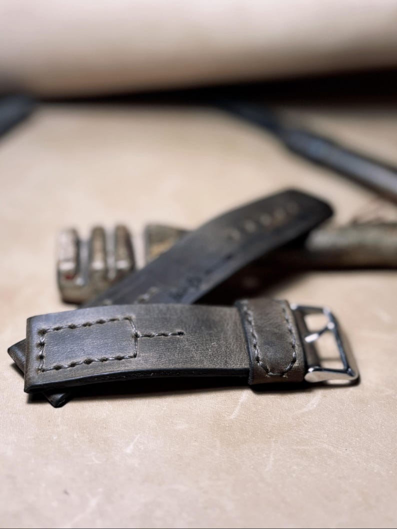 May include: Close-up of a grey leather watch strap with a silver buckle. The strap has visible stitching and a rectangular loop. Another strap is visible in the background, along with a metal tool.