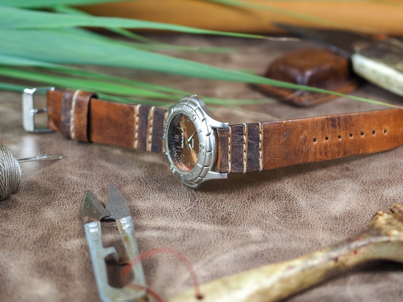 May include: A brown leather watch strap with white stitching. The strap is attached to a silver watch with a round face.