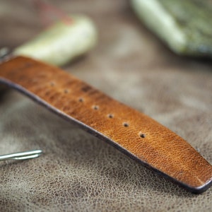 May include: A close-up of a brown leather watch strap with stitching and multiple holes for adjusting the size.