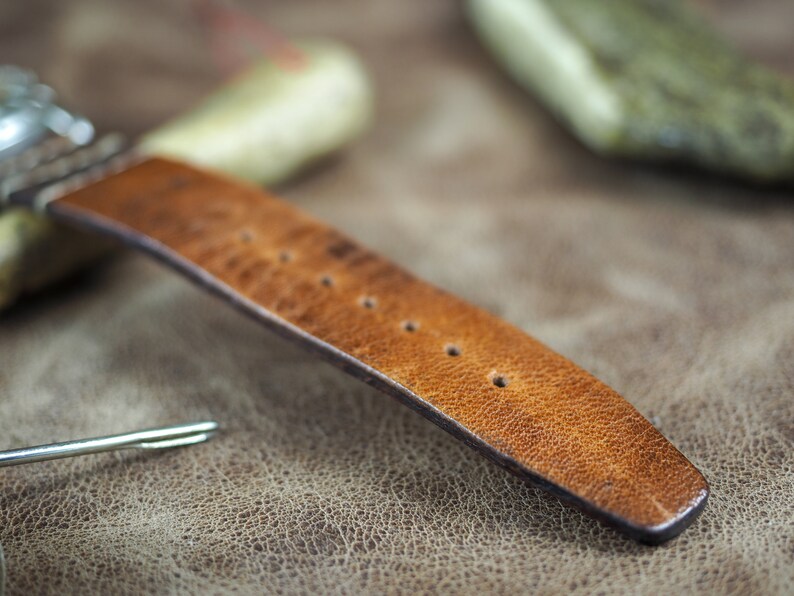 May include: A close-up of a brown leather watch strap with stitching and multiple holes for adjusting the size.