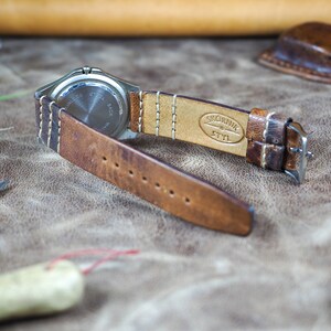 May include: A brown leather watch strap with white stitching. The strap is attached to a silver watch with a stainless steel back. The strap is embossed with the text "SKORNIK & STYL".