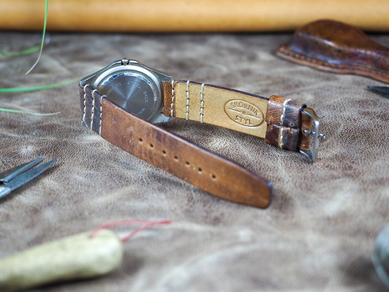 May include: A brown leather watch strap with white stitching. The strap is attached to a silver watch with a stainless steel back. The strap is embossed with the text "SKORNIK & STYL".