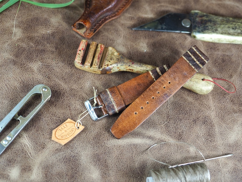 May include: A brown leather watch strap with white stitching and a silver buckle. The strap is lying on a brown leather surface with other leatherworking tools.