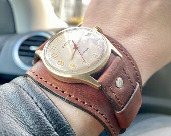 May include: A gold-toned wristwatch with a brown leather band. The watch face has a white background with black Roman numeral hour markers and black hands. The watch face also has the text "Wostok" and "18 Jewels".