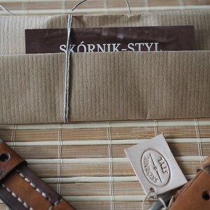 May include: A brown leather watch strap with white stitching and a brown paper package tied with twine. The package has a business card that says "SK&Oacute;RNIK-STYL" and a white leather tag with the text "SK&Oacute;RNIK-STYL" in a circle.