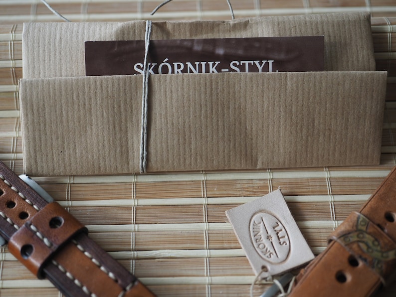 May include: A brown leather watch strap with white stitching and a brown paper package tied with twine. The package has a business card that says "SK&Oacute;RNIK-STYL" and a white leather tag with the text "SK&Oacute;RNIK-STYL" in a circle.