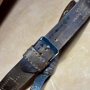 May include: Close-up of a brown leather watch strap with a silver buckle. The strap has decorative stitching and etched symbols. The leather shows wear and a vintage aesthetic. The strap is likely for a watch.
