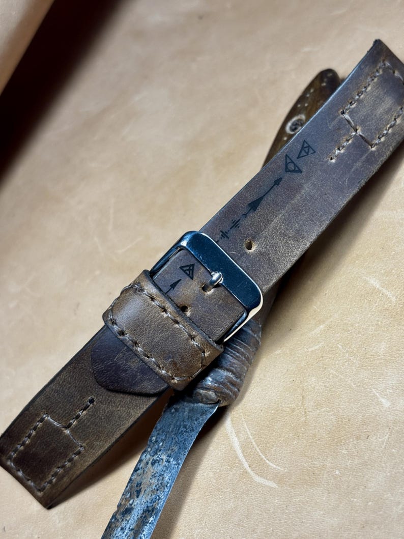 May include: Close-up of a brown leather watch strap with a silver buckle. The strap has decorative stitching and etched symbols. The leather shows wear and a vintage aesthetic. The strap is likely for a watch.
