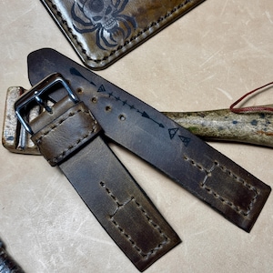 May include: Close-up of a brown leather watch strap with a silver buckle. The strap has decorative stitching and arrow designs. A matching leather wallet with a skull spider design is in the background.