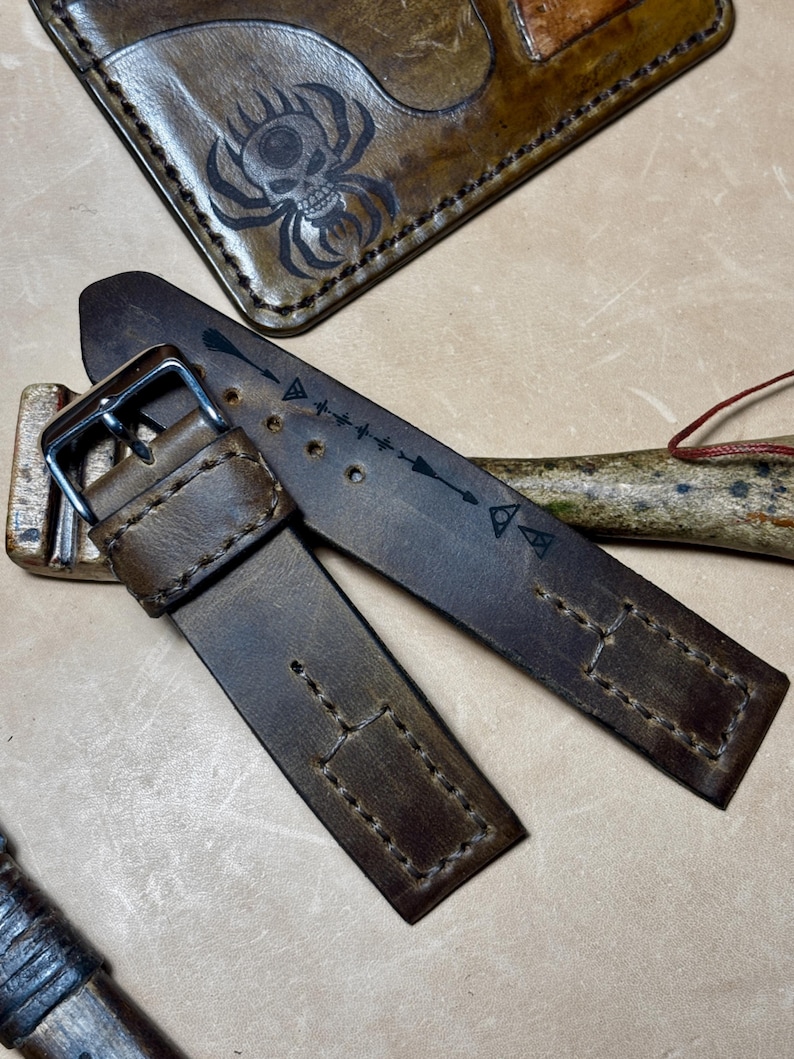 May include: Close-up of a brown leather watch strap with a silver buckle. The strap has decorative stitching and arrow designs. A matching leather wallet with a skull spider design is in the background.