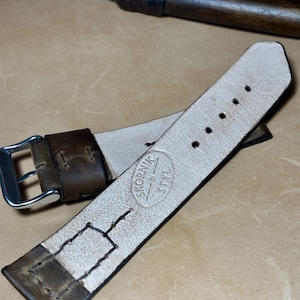 May include: A brown leather watch strap with a silver buckle. The strap has a light-coloured interior and is stamped with the text "SKORNIK STYL". A knife with a wooden handle is in the background.