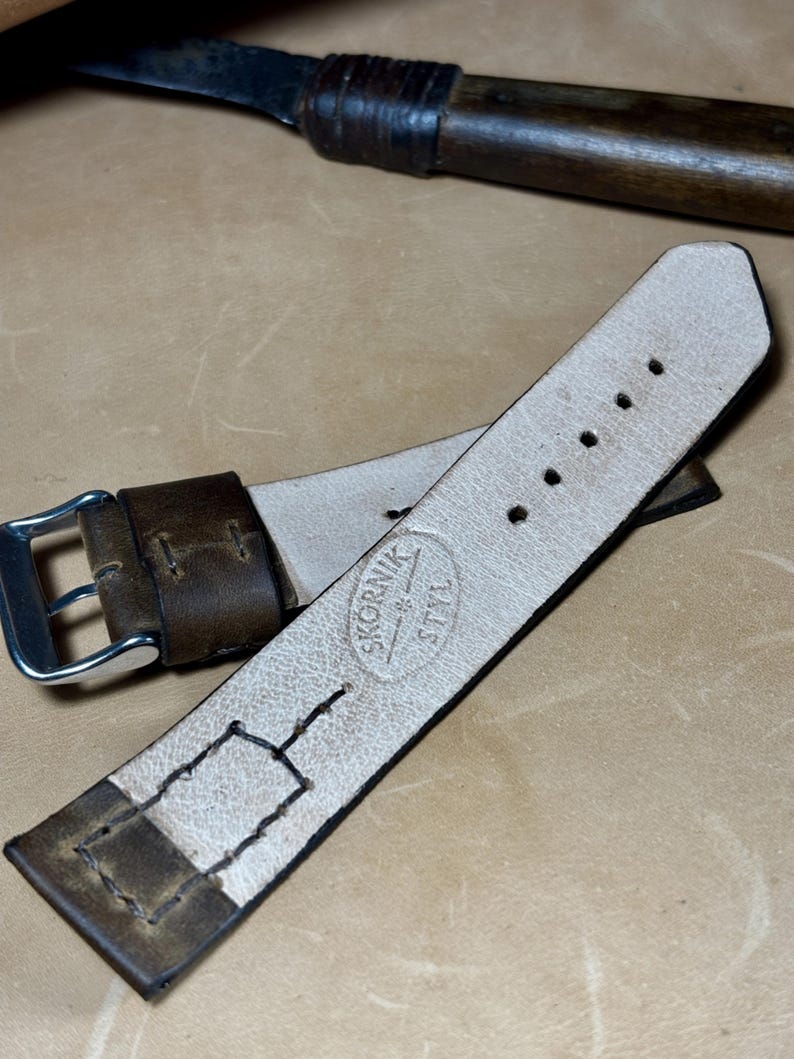May include: A brown leather watch strap with a silver buckle. The strap has a light-coloured interior and is stamped with the text "SKORNIK STYL". A knife with a wooden handle is in the background.