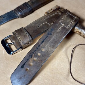 May include: A close-up of a distressed brown leather watch strap with a silver buckle. The strap features decorative stitching and arrow symbols. A vintage knife and a needle with thread are also visible.
