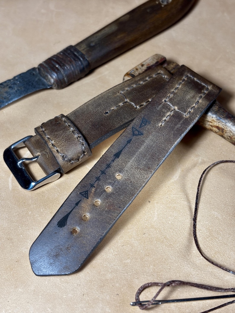May include: A close-up of a distressed brown leather watch strap with a silver buckle. The strap features decorative stitching and arrow symbols. A vintage knife and a needle with thread are also visible.
