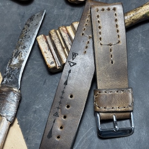 Op de afbeelding: Een bruine leren horlogeband met een zilveren gesp. De band heeft decoratieve stiksels en geëtste pijl- en driehoekontwerpen. De horlogeband wordt getoond met een mes en andere lederen accessoires.