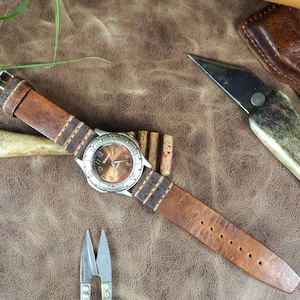 May include: A brown leather watch strap with white stitching and a silver watch face with a brown background. The watch is on a brown leather surface with other leatherworking tools.