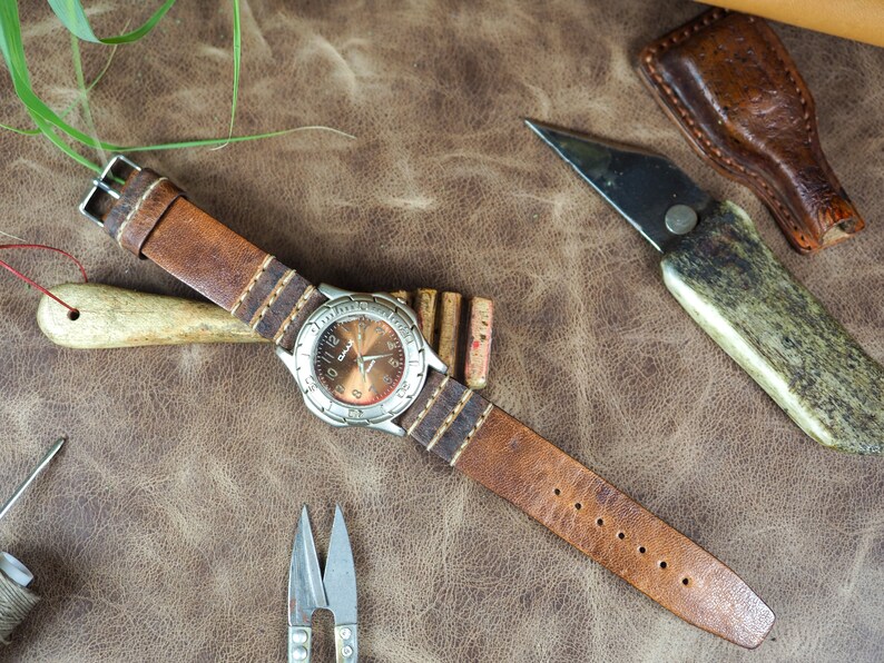 May include: A brown leather watch strap with white stitching and a silver watch face with a brown background. The watch is on a brown leather surface with other leatherworking tools.