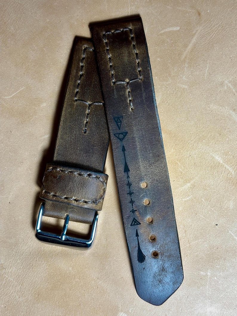 May include: Two brown leather watch straps with white stitching and a silver buckle. One strap features a series of black symbols and arrows. The leather has a distressed appearance.
