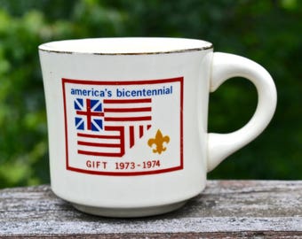 Puede incluir: Taza de cerámica color crema con borde dorado y asa blanca. La taza presenta un gráfico rojo, blanco y azul que conmemora el bicentenario de Estados Unidos, con el texto "america's bicentennial" y "GIFT 1973-1974".