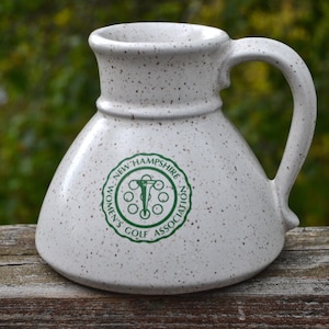 Puede incluir: Jarra de cerámica blanca moteada con asa, con un emblema verde que dice "New Hampshire Women's Golf Association". La jarra tiene una base ancha y un cuello estrecho, perfecta para servir bebidas. Mide unos 15 cm de alto.
