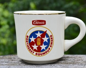 Puede incluir: Taza de cerámica blanca con borde dorado y asa grande. La taza presenta el logotipo de "Gino's" y un emblema circular con estrellas y el texto "Northeast Roundup Award". La taza está sobre un fondo verde borroso.