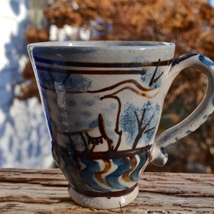May include: A handmade ceramic mug with a unique design. The mug features a light blue and white base with brown and blue painted details, including trees and abstract shapes. The handle is also decorated with blue accents.