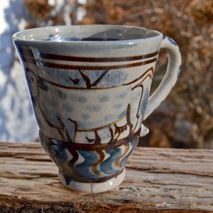 May include: A handmade ceramic mug with a unique design. The mug features a light blue and white base with brown and blue painted details. The handle is white. The mug is sitting on a wooden surface.