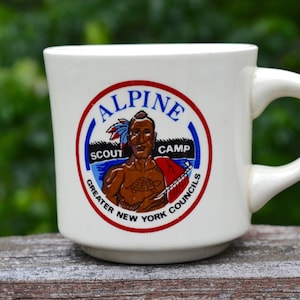 Op de afbeelding: Witte keramische mok met een cirkelvormig embleem met de tekst "ALPINE SCOUT CAMP GREATER NEW YORK COUNCILS". Het embleem toont een Indiaanse figuur. De mok heeft een gebogen handvat en staat tegen een vage groene achtergrond.