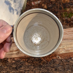 May include: A handmade ceramic mug with a speckled, off-white glaze and a dark brown rim. The mug is held by a hand, showcasing its interior and handle. The mug is sitting on a piece of wood.
