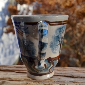 May include: Handmade ceramic mug with a unique design. The mug features a light blue and brown color scheme with a landscape design. The handle is striped with blue and white. The mug is approximately 4 inches tall.