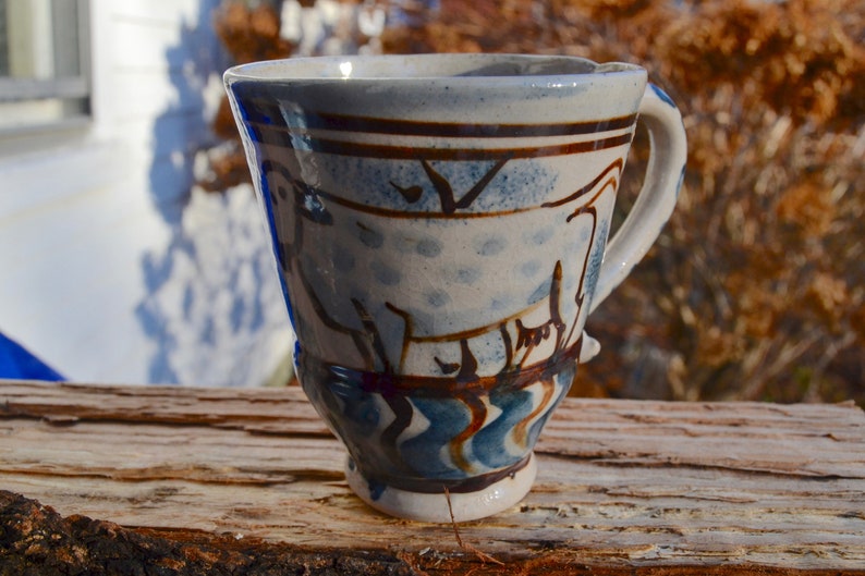 May include: Handmade ceramic mug with a unique design. The mug features a white base with blue and brown painted details, including abstract shapes and patterns. The handle is curved and the mug sits on a wooden surface.