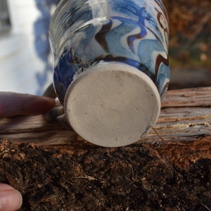 May include: A handmade ceramic mug with a blue and brown abstract design. The mug has a white base and a handle. The mug is resting on a piece of wood.