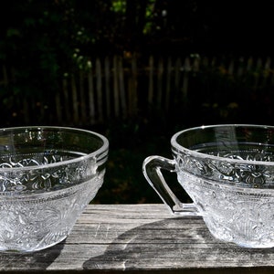 May include: Two clear glass teacups with handles. The cups have a detailed floral pattern around the middle and are sitting on a wooden surface. The background is a blurred view of a fence and trees.