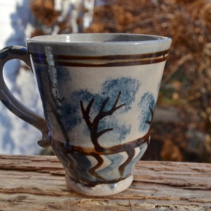 May include: Handmade ceramic mug with a blue and brown tree design. The mug has a curved handle and a flared base. The exterior features a light background with blue tree silhouettes and brown accents. The mug is approximately 4 inches tall.