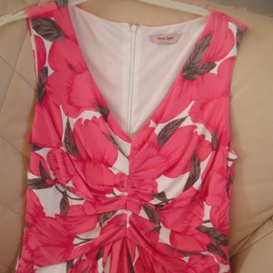 Peut inclure: Une robe sans manches avec un décolleté en V et un imprimé floral. La robe est blanche avec des fleurs roses et des feuilles brunes. La robe a une fermeture éclair dans le dos et une étiquette qui dit "Next Style".