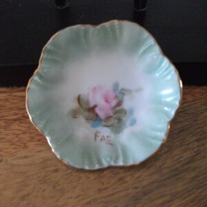 Peut inclure: Un petit plat en porcelaine vert et blanc à bords festonnés et bordure dorée. Le plat est décoré d'une rose rose peinte à la main avec des feuilles vertes et des accents bleus. Le mot "Fae" est écrit en lettres dorées au bas du plat.