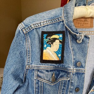 May include: A blue denim jacket with a black patch featuring a woman in a yellow and blue kimono. The patch is on the left side of the jacket, above the pocket. The jacket has a Calvin Klein label on the back of the collar.