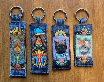May include: Four denim keychains with silver key rings. Each keychain features a unique design: one with a colorful floral pattern, two with cat portraits adorned with floral crowns, and one with a blue and yellow floral design. The keychains are on a wooden surface.