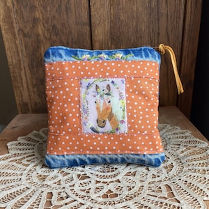 May include: A small, square pouch with a horse portrait. The pouch features a denim blue top and bottom, with an orange polka-dot center panel. The horse portrait is framed by a floral wreath. A yellow leather strap is attached to the side.