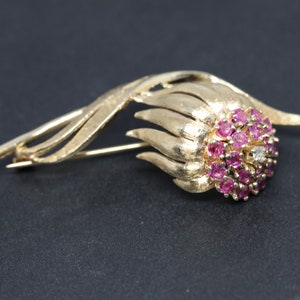 May include: A gold brooch featuring a stylised flower design. The flower is crafted from gold petals and has a centre of pink and white stones.