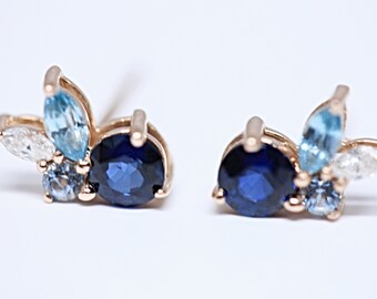 May include: A pair of gold earrings with blue and white gemstones. The earrings feature a cluster of small blue and white gemstones surrounding a larger blue gemstone.