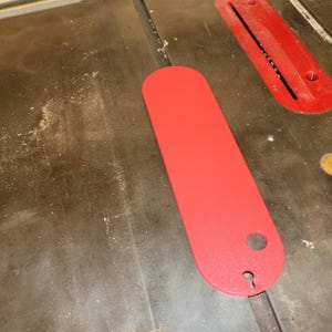 May include: A red table saw throat plate and a red insert plate are on a wooden table saw surface. The throat plate is long and oval-shaped with a hole at one end. The insert plate is rectangular with a slot.