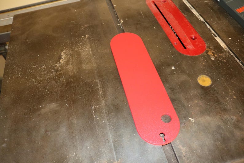 May include: A red table saw throat plate and a red insert plate are on a wooden table saw surface. The throat plate is long and oval-shaped with a hole at one end. The insert plate is rectangular with a slot.