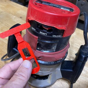 May include: A red and black router with a red plastic key. The router has a depth gauge and a lock/unlock switch. The key has a T-shaped handle and a slot for the router shaft.
