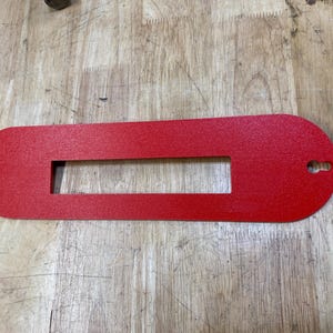 May include: A red, oval-shaped, metal template with a rectangular cutout in the center. The template has a small hole in the top right corner.